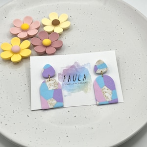 Op de afbeelding: Een paar pastelkleurige geometrische oorbellen in paars, blauw en wit met gouden accenten. De oorbellen worden gepresenteerd op een witte kaart met de tekst "PAULA JEWELLERY DESIGN".