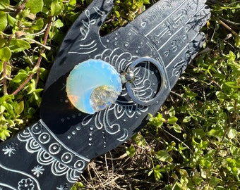 May include: A silver key chain with a white opal stone attached. The stone has a rainbow iridescent sheen. The key chain is on a black hand-shaped background.