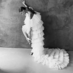 Op de afbeelding: Een zwart-wit foto van een witte gelaagde jurk met een lange sleep. De jurk heeft een gerimpeld lijfje en rok. De persoon draagt ​​hoge hakken. De achtergrond is een getextureerde grijze muur en witte vloer.