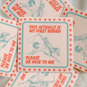 May include: A pink and blue square sticker with a cowboy riding a bucking horse. The sticker has a rope lasso and the text "This Actually Is My First Rodeo" and "Please Be Nice To Me".