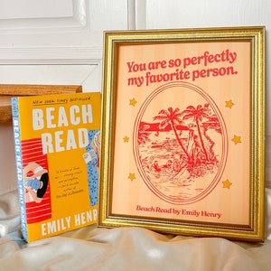 May include: A framed print with a peach background and red illustration of palm trees and people in a lake. The text "You are so perfectly my favorite person." is at the top of the print. The text "Beach Read by Emily Henry" is at the bottom of the print. A book titled "Beach Read" by Emily Henry is in the foreground.