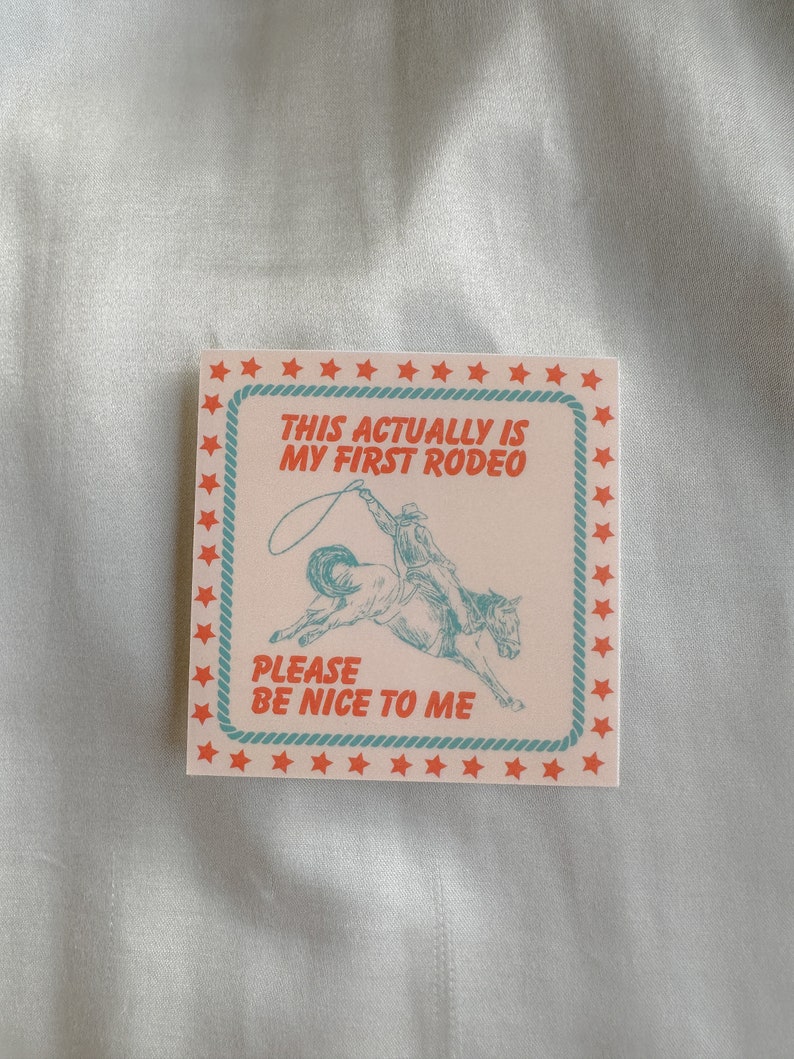 May include: A light pink card with a blue and white illustration of a cowboy riding a bucking horse. The card has a red border with white stars and the text "This actually is my first rodeo. Please be nice to me."