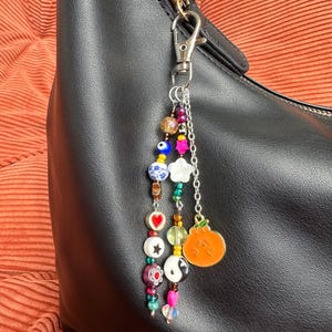 May include: A beaded charm with a lobster clasp attached to a black handbag. The charm features a variety of colorful beads, including a heart, a flower, and a pumpkin charm. The beads are strung together with a silver chain.