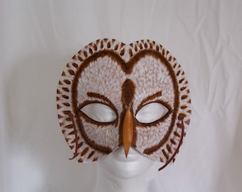 May include: A handcrafted owl mask featuring a white and brown feathered pattern with a pointed orange beak. The mask has two large eye openings and brown leather ties on the sides for securing it. The design mimics the facial features of an owl.