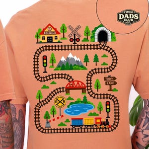 May include: Orange t-shirt with a colorful train track design. The graphic includes a train, tunnel, bridge, lake, and the words "Tired Dads Club". The design is a playful illustration of a train journey.