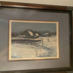 May include: A framed winter landscape painting depicting three wooden houses in a snowy field. The houses are brown and have white snow on their roofs. The field is covered in white snow and there are trees in the background. The painting is framed in a dark wood frame.