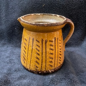 May include: A brown ceramic pitcher with a wide mouth and a handle. The pitcher has a brown and yellow glaze with a geometric pattern.