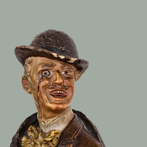 Puede incluir: Una figurita de cerámica de un hombre con un sombrero bombín marrón y una chaqueta marrón. Tiene bigote y un monóculo.