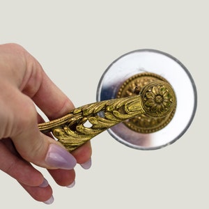 Könnte beinhalten: Ein verzierter, goldfarbener Dekor-Griff mit einem floralen Muster und einem runden, dekorativen Knopf. Der Griff ist an einer runden, silberfarbenen Basis befestigt.