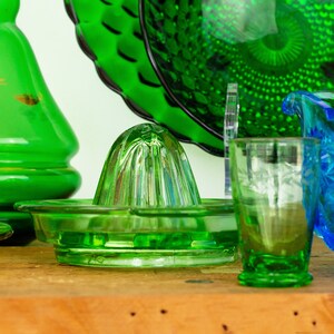 以下が含まれることがあります： 木製の表面に、溝のあるデザインの緑色のガラス製の柑橘類ジューサーが置かれています。背景には、緑色のガラス製の飲み物グラスと青いガラス製のピッチャーも見えます。