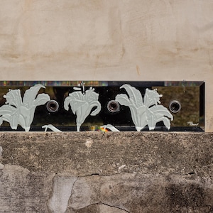 May include: A rectangular wall-mounted mirror fixture featuring a dark reflective surface with three circular light sockets. The glass is decorated with etched white lily floral patterns across the front, and the signature F. Ramond is visible.