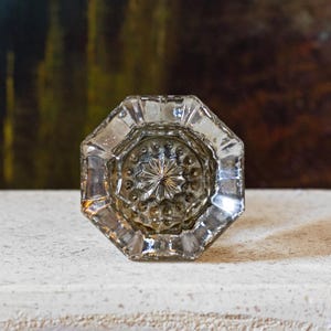 May include: A clear, octagonal glass door knob featuring a central starburst pattern surrounded by a ring of small, raised circular beads. The glass has a faceted edge design that reflects light, creating a multi-dimensional appearance.