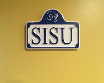 May include: A white sign with the word "SISU" in bold, blue letters. The sign has a blue border and a decorative top with floral scrollwork. The sign is mounted on a yellow wall.