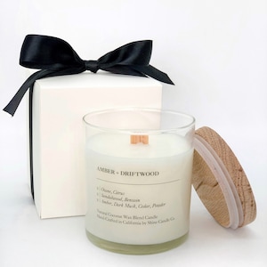 May include: A white candle in a glass jar with a wooden lid. The candle is labelled "Amber + Driftwood" and lists the scent notes: Ozone, Citrus, Sandalwood, Benzoin, Amber, Dark Musk, Cedar, Powder. The label also states "Natural Coconut Wax Blend Candle" and "Hand-Crafted in California by Shine Candle Co."