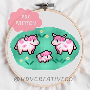 Puede incluir: Un patrón de punto de cruz en un bastidor de bordado de madera. El diseño presenta tres vacas rosas y blancas pixeladas sobre un campo verde con pequeñas flores blancas. El texto "PDF PATTERN" está en forma de nube rosa. El nombre de la tienda es visible en la parte inferior.