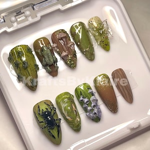 Könnte beinhalten: Ein Set mit zehn grünen und braunen falschen Nägeln mit verschiedenen Designs zum Thema Natur, darunter ein Frosch, Pilze, eine Spinne und Blumen. Die Nägel sind in einer weißen Box angeordnet.