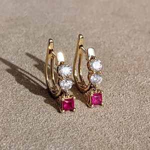 May include: A pair of gold hoop earrings featuring two clear round stones set vertically on the front of each hoop, with a single square-cut red stone dangling from the bottom of each earring, creating a symmetrical and elegant jewelry design.
