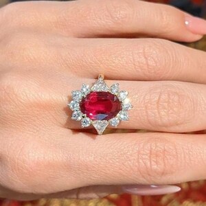 K&ouml;nnte beinhalten: Ein Ring mit einem gro&szlig;en, oval geschliffenen, tiefroten Edelstein in der Mitte, umgeben von einem Kranz aus zw&ouml;lf kleinen, klaren, rund geschliffenen wei&szlig;en Steinen in einer Metallfassung, die ein bl&uuml;tenartiges Design bilden.