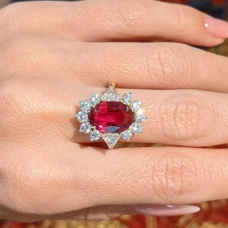 K&ouml;nnte beinhalten: Ein Ring mit einem gro&szlig;en, oval geschliffenen, tiefroten Edelstein in der Mitte, umgeben von einem Kranz aus zw&ouml;lf kleinen, klaren, rund geschliffenen wei&szlig;en Steinen in einer Metallfassung, die ein bl&uuml;tenartiges Design bilden.