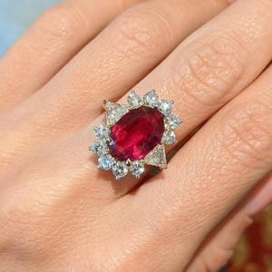 K&ouml;nnte beinhalten: Ein Ring mit einem gro&szlig;en, oval geschliffenen, tiefroten Edelstein in der Mitte, umgeben von einem Kranz aus kleineren, rund geschliffenen wei&szlig;en Diamanten und zwei dreieckig geschliffenen wei&szlig;en Diamanten an den Seiten, gefasst in Metall.