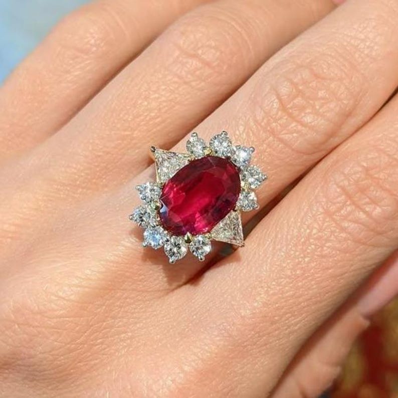 K&ouml;nnte beinhalten: Ein Ring mit einem gro&szlig;en, oval geschliffenen, tiefroten Edelstein in der Mitte, umgeben von einem Kranz aus kleineren, rund geschliffenen wei&szlig;en Diamanten und zwei dreieckig geschliffenen wei&szlig;en Diamanten an den Seiten, gefasst in Metall.