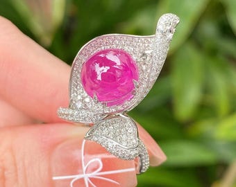 Peut inclure: Une bague argentée ornée d'une grande pierre précieuse rose vif taillée en cabochon rond au centre. La bague a la forme d'une feuille ou d'un pétale stylisé, avec toute la surface incrustée de nombreuses petites pierres blanches étincelantes.