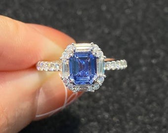 Peut inclure: Une bague en argent ornée d'une grande pierre précieuse rectangulaire bleu foncé, entourée de petites pierres précieuses claires. La bande de la bague est également ornée de petites pierres précieuses claires, créant un effet scintillant. La bague est posée sur un fond sombre.