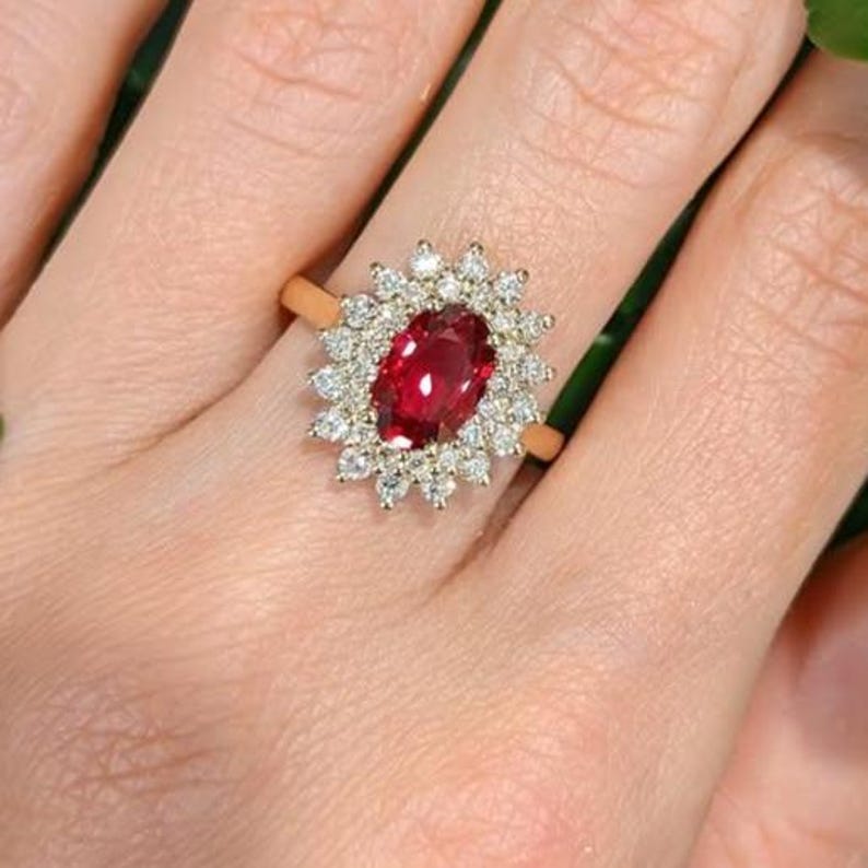 May include: A gold ring featuring a central oval-cut deep red ruby surrounded by a halo of small, round, clear white diamonds. The gemstone setting is intricate, with the diamonds arranged in a floral-like pattern around the ruby centre.