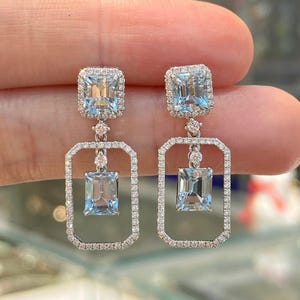 Puede incluir: Un par de elegantes pendientes colgantes con piedras preciosas rectangulares de color azul claro. Cada pendiente tiene una parte superior cuadrada con un halo de pequeñas piedras transparentes, conectada a un marco rectangular con una piedra preciosa a juego en la parte inferior.