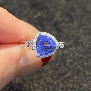 Puede incluir: Un anillo de plata con una gran piedra preciosa triangular de color azul intenso, rodeada por un halo de piedras claras más pequeñas. Piedras claras adicionales están engastadas a ambos lados de la piedra principal. El anillo es sostenido por dedos.
