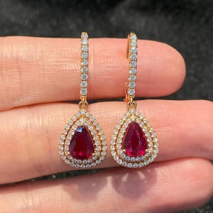 May include: A pair of drop earrings featuring pear-shaped red rubies surrounded by a double halo of small white diamonds. The earrings are set in yellow gold with a vertical bar of diamonds extending upward from the central ruby setting.