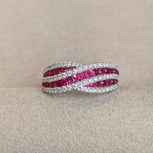May include: A crossover band ring featuring two intertwined rows of round-cut vibrant red rubies and two rows of sparkling white diamonds set in a polished metal band. The design creates a continuous woven pattern with alternating gemstone rows.