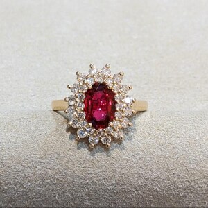 May include: A gold ring featuring a central oval-cut deep red gemstone surrounded by a halo of small, round, clear white stones. The setting is intricate, with the clear stones arranged in a sunburst pattern around the central red stone.