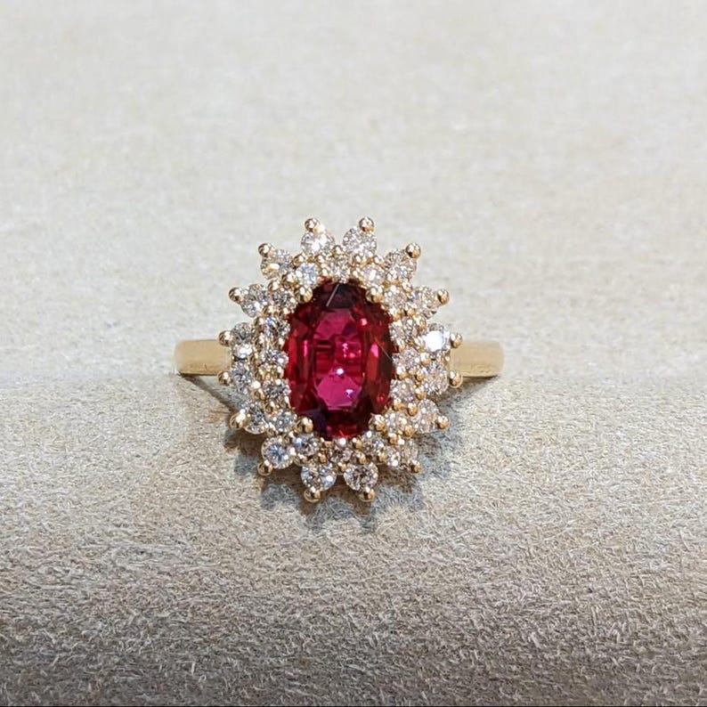 May include: A gold ring featuring a central oval-cut deep red gemstone surrounded by a halo of small, round, clear white stones. The setting is intricate, with the clear stones arranged in a sunburst pattern around the central red stone.