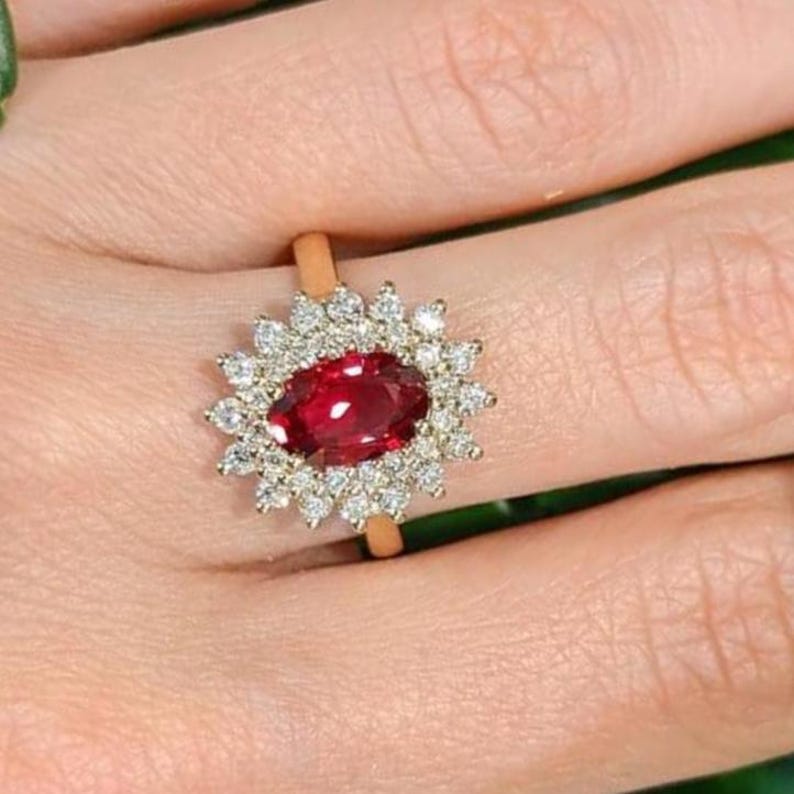 May include: A gold ring featuring a central oval-cut red ruby surrounded by a halo of small, round, clear white diamonds. The gemstone setting is intricate, with the diamonds arranged in a sunburst pattern around the deep red centre stone.
