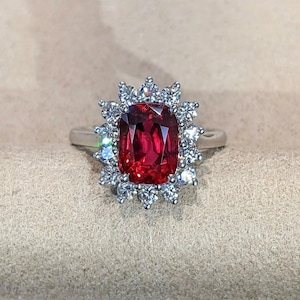 Op de afbeelding: Een zilverkleurige ring met een grote, rechthoekige, dieprode edelsteen in het midden. De centrale steen is omringd door een halo van talloze kleine, heldere, rondgeslepen stenen in een stralend patroon, op een gladde, gepolijste metalen ring.