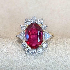 K&ouml;nnte beinhalten: Ein Goldring mit einem gro&szlig;en, oval geschliffenen roten Rubin in der Mitte. Der Rubin ist von einem Kranz aus zehn runden Brillanten und zwei gr&ouml;&szlig;eren dreieckig geschliffenen Diamanten an den Seiten umgeben, gefasst in eine Ringschiene aus Gelbgold.
