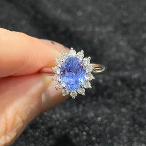 Puede incluir: Un anillo de plata con una gran piedra preciosa azul ovalada, rodeada por un halo de piedras claras más pequeñas. El diseño del anillo es elegante, con un enfoque en la piedra azul central.