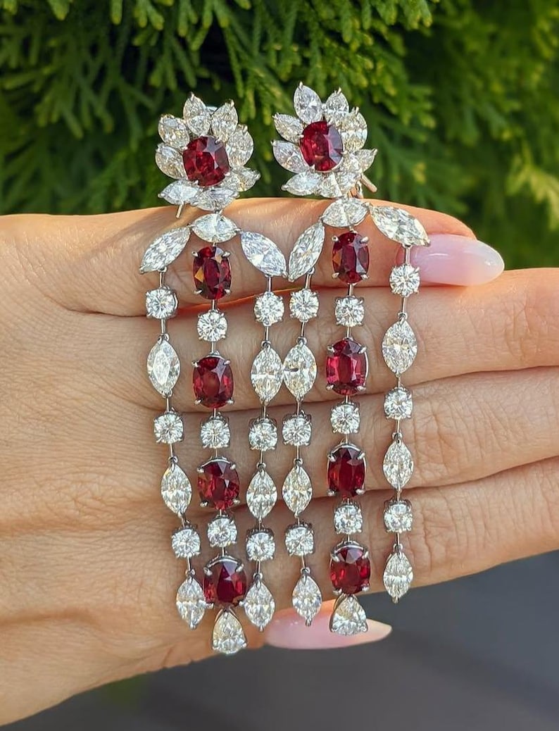 Puede incluir: Un par de pendientes largos colgantes con rub&iacute;es ovalados de color rojo intenso y diamantes transparentes en talla marquesa y redonda. El dise&ntilde;o incluye un motivo floral superior con un rub&iacute; central, seguido de varias cadenas verticales que alternan rub&iacute;es y diamantes.