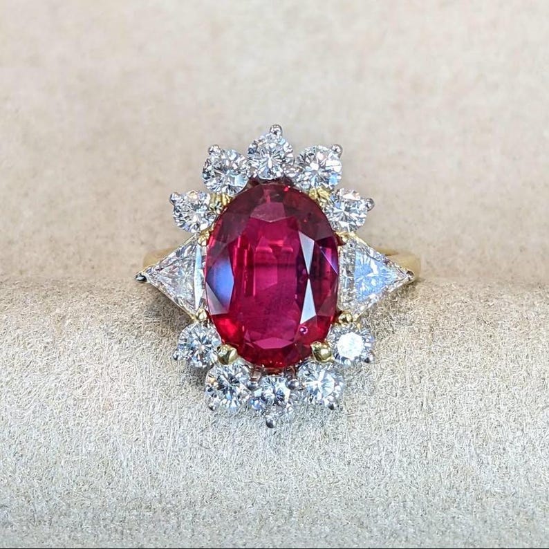 K&ouml;nnte beinhalten: Ein Goldring mit einem gro&szlig;en, oval geschliffenen, tiefroten Rubin in der Mitte, umgeben von einem Kranz aus runden Brillanten und zwei dreieckig geschliffenen Diamanten an den Seiten, gefasst in eine Metallschiene mit poliertem Finish und Krappen.