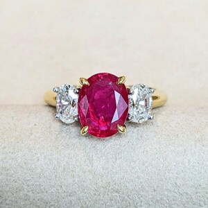 May include: A three-stone ring featuring a large oval-cut vibrant red ruby center stone set in yellow gold prongs, flanked by two oval-cut clear white diamonds set in white gold prongs on a polished yellow gold band.