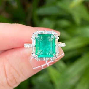 Puede incluir: Un anillo de tono plateado con una gran piedra preciosa verde rectangular de corte esmeralda en el centro. La piedra está rodeada por un halo de pequeños diamantes transparentes de corte baguette y redondo, con diamantes adicionales en el aro.