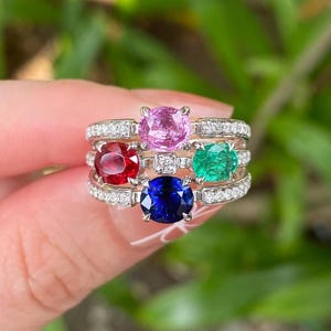 Könnte beinhalten: Eine Sammlung von vier Edelsteinringen. Jeder Ring hat einen andersfarbigen Edelstein: rosa, rot, grün und blau. Die Ringe sind in einem goldenen Band mit kleinen, klaren Akzentsteinen gefasst. Die Ringe sind aufeinander gestapelt.