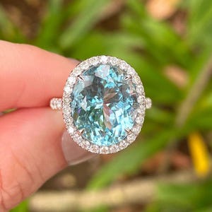 Puede incluir: Un anillo con una piedra preciosa de aguamarina de corte ovalado, de color azul claro. La piedra central está rodeada por un halo de pequeños diamantes transparentes. La banda del anillo es de metal claro, probablemente plata u oro blanco. Una joya.
