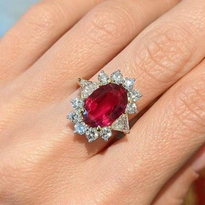K&ouml;nnte beinhalten: Ein Ring mit einem gro&szlig;en, oval geschliffenen, tiefroten Rubin als Mittelstein. Der Rubin ist von einer Gruppe kleinerer, rund geschliffener wei&szlig;er Diamanten und zwei gr&ouml;&szlig;eren, dreieckig geschliffenen wei&szlig;en Diamanten an den Seiten umgeben.