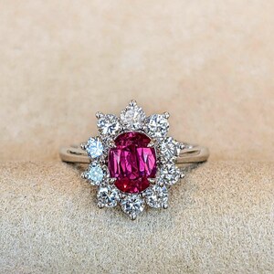 May include: A silver-toned ring featuring a central oval-cut deep pink gemstone surrounded by a halo of ten round-cut clear white stones. The metal band is smooth and polished, supporting the floral-shaped cluster setting of the ring.