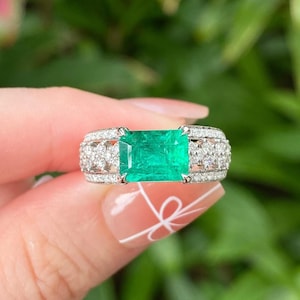 May include: A silver-toned ring featuring a large, rectangular, vibrant green emerald gemstone at the center, flanked by two sections of multiple small, clear, round-cut diamonds set into the band, creating a wide, multi-faceted metallic appearance.