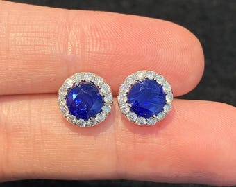 Peut inclure: Une paire de boucles d'oreilles à clous rondes présentant une pierre précieuse circulaire d'un bleu profond et vibrant au centre. Chaque pierre centrale est entourée d'un halo de petites pierres blanches claires et étincelantes sur une monture en métal argenté.
