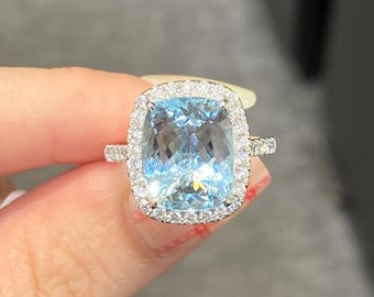 Peut inclure: Une bague ornée d'une grande pierre précieuse bleu clair taille coussin, montée sur un anneau en métal argenté. La pierre centrale est entourée d'un halo de petites pierres claires taille ronde, avec d'autres pierres incrustées sur les côtés.
