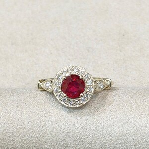 Pode incluir: Um anel de ouro com uma pedra central de rubi vermelho profundo de corte redondo, rodeada por um halo de pequenos diamantes transparentes. A banda apresenta detalhes metálicos decorativos com diamantes adicionais cravados nas laterais.